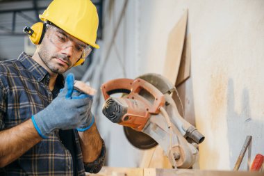 Marangoz adam sarı kasketli, güvenlik gözlüklü arka planda dairesel testere ile odun kesmek için ölçü alıyor. Marangozluk ve marangozluk atölyesi ayarları için ideal, Ulusal Marangozlar Günü