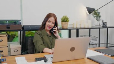 Happy Asian young woman working on laptop computer at home office while talking on mobile phone, smiling businesswoman having business call to talking with company sales client financial