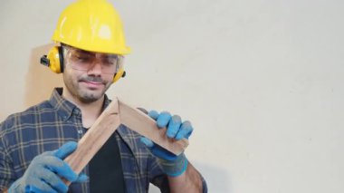 Sarı kasketli, güvenlik gözlüklü ve mavi eldivenli inşaat işçisi marangozluk atölyesindeki bir tahta parçasını inceliyor. Ahşap işçiliği, işçiliği ve kalite kontrolü, Ulusal Marangozlar Günü