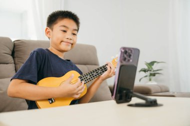 Genç bir çocuk bir kanepede oturup ukulele çalıyor ve bir standa monte edilmiş akıllı bir telefonla online müzik dersi veriyor. Konforlu ev ayarları etkileşimli öğrenme ve yaratıcı ifadeyi vurgular.