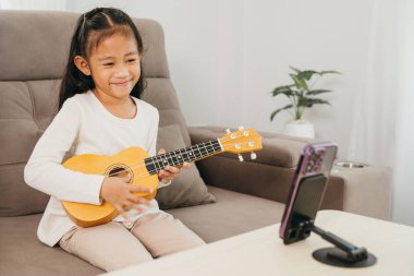 Akıllı bir telefon seansını kaydederken Asyalı bir kız gitar tıngırdatıyor. Ev tabanlı düzende rahat bir ortamda dijital öğrenme ve yaratıcılık vurgulanıyor.