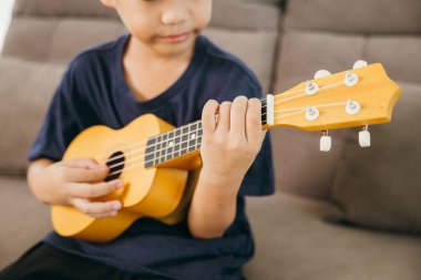 Kanepede oturmuş, ukulele çalan Asyalı bir çocuğun portresi. Oturma odası, enstrümanların detaylarını vurgulayan sıcak ve yaratıcı bir ortam sağlıyor..