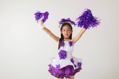 Ponpon kız gibi giyinmiş, elinde mor ponponlar tutan neşeli bir genç kız. Temiz beyaz bir arkaplanla eğlenceli bir stüdyoda çekildi. Amigo kız.