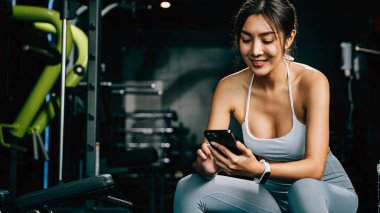 Asyalı kadın yedek kulübesinde dinleniyor ve beden eğitimi aldıktan sonra tüm güncellemeleri kontrol ediyor. Kadın spor salonunda spor yaparken cep telefonu uygulaması kullanıyor. Sağlıklı yaşam tarzı konsepti.