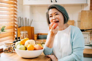 Evdeki mutfakta, yaşlı bir Asyalı kadın, bir büyükanne, sağlıklı vejetaryen yemekleri pişirip hazırlarken eğleniyor. Taze elma seçimi diyet konseptini ve mutluluğunu yansıtıyor..