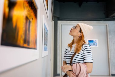 Asyalı kadın renkli çerçeveli resimlerin önünde dikilip beyaz duvara bakıyor, insanlar fotoğraf çerçevesinde sergi sanat galerisine yaslanıyor,