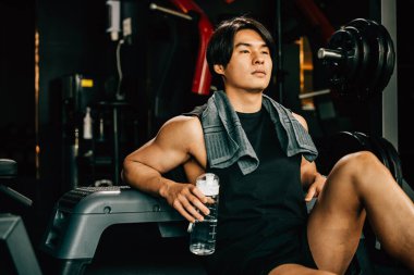 Spor kıyafetleriyle eğitilmiş Asyalı bir gencin portresi spor salonunda antrenman, ağırlık eğitimi, sağlıklı yaşam tarzı konsepti sonrasında elinde su ya da proteinle bankta dinleniyor.