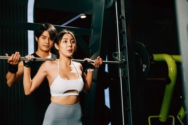 Asyalı kadın, spor salonundaki antrenörün yardımıyla ağırlık kaldıran spor salonundaki erkek, spor salonundaki kadın halterciye halter kaldırarak antrenman yapıyor.