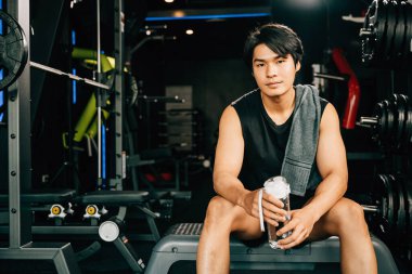 Asyalı genç adam portresi siyah spor kıyafetleri içinde dinlenmek için oturmuş spor salonunda su ya da protein tutarken ağırlık antrenmanı, sağlıklı yaşam tarzı konsepti.