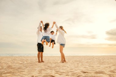 Sahilde bir Asyalı ailenin çocukları kucağında tutması sevinçle havaya zıplıyor. Güzel okyanus arka planı aile mutluluğunun ve kaygısız eğlencenin özünü yakalar..
