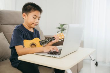 Oturma odasındaki bir kanepede oturan bir çocuk, dizüstü bilgisayarındaki ukulele dersini takip ediyor. Sahne modern internet öğrenimini vurguluyor. Müziği evinde rahat bir şekilde çalışıyor..