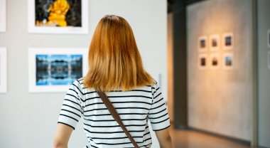 Asian young woman standing she looking art gallery in front of colorful framed paintings pictures on white wall, female watch at photo frame to leaning against at exhibit museum, Back view