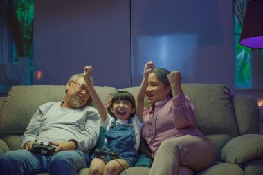 Konforlu bir koltukta video oyunu oynarken mutlu Asyalı aile bağı. Küçük kız, kanepede otururken büyük anne ve babasıyla bilgisayar oyunu konsolu oynadıktan sonra kazanıyor.