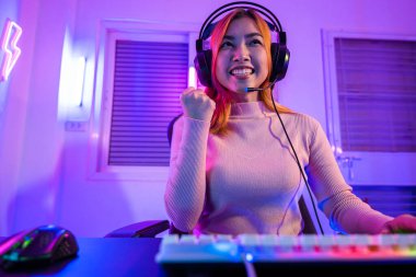 Zafer kazanmak. Asyalı oyuncu online video oyunu oynuyor. Masaüstü bilgisayarında geceleri mor ışık yanıyor. Oyun kulaklıklı mutlu kadın başarılı olduğu bilgisayarı kullanıyor, E-Sport kavramı.