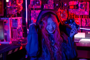 Happy Gamer sanal turnuva için kumanda kolu tutuyor bilgisayarla çevrimiçi video oyunu oynuyor kutlama yapmak için elini kaldırıyor oyun kulaklığı takan Asyalı kadın heyecanlanıyor ve kazanıyor.