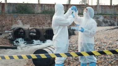 Nehirden gelen suyu test tüpü, koruyucu giysi içindeki bilim adamı veya biyolog ile örnekleyen ve maskesini koruyan ekolojist, analiz için endüstriden atık su örneği topluyor, sorun ortamı