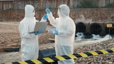 Nehirdeki suyu test tüpüyle örnekleyen ve dizüstü bilgisayara veri kaydeden bir ekolojist koruyucu giysi içindeki biyolog endüstriyel ve sorunlu ortamdan atık su örneği topluyor.