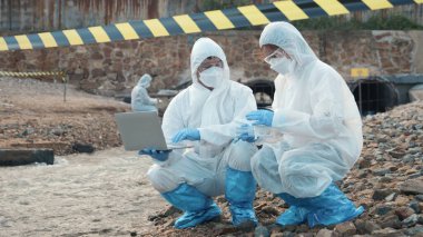 Nehirdeki suyu test tüpüyle örnekleyen ve dizüstü bilgisayara veri kaydeden bir ekolojist koruyucu giysi içindeki biyolog endüstriyel ve sorunlu ortamdan atık su örneği topluyor.