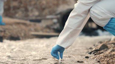 Nehirdeki suyu test tüpüyle örnekleyen ekolojist, koruyucu kıyafet giyiyor ve maske endüstriyel, sorunlu çevreden ve bilim adamı çevreciden atık su örnekleri topluyor.