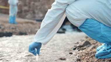 Nehirdeki suyu test tüpüyle örnekleyen ekolojist, koruyucu kıyafet giyiyor ve maske endüstriyel, sorunlu çevreden ve bilim adamı çevreciden atık su örnekleri topluyor.