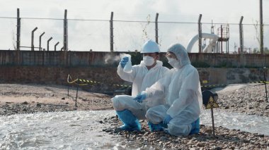 Nehirden çıkan zehirli kimyasalları test tüpü camlarıyla test eden ve beyaz duman çıkaran ekolojist, koruyucu kıyafet giyiyor ve maske endüstriyel, sorunlu ortamdan örnek su topluyor.