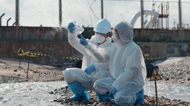 Ecologist sampling water of toxic chemicals from river touching screen on digital tablet, Biologist wear protective suit and mask collects sample of waste water from industrial, problem environment