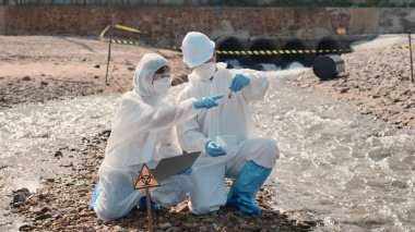 Biyolog koruyucu kıyafet giyer ve maske endüstriden, sorunlu çevreden atık su numunesi toplar, ekolojist örneği incelemek ve veri kaydetmek için ölü balığı laptopa, toksik suya götürür.