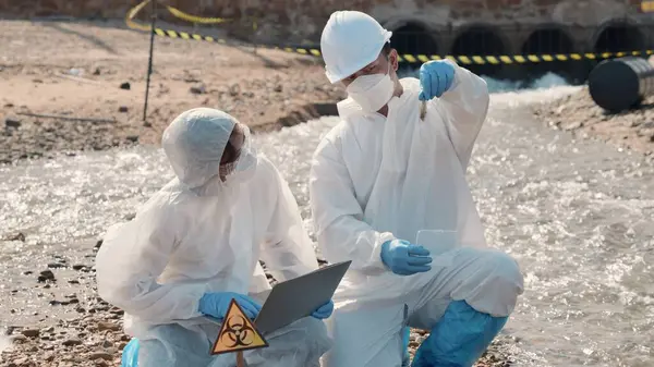 Biyolog koruyucu kıyafet giyer ve maske endüstriden, sorunlu çevreden atık su numunesi toplar, ekolojist örneği incelemek ve veri kaydetmek için ölü balığı laptopa, toksik suya götürür.