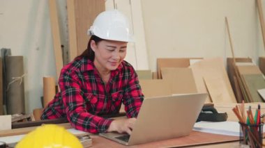 Beyaz kasketli ve kırmızı ekose gömlekli marangoz kadın etrafı aletler, tahta kırıntıları ve kalemlerle çevrili bir marangoz dükkanında dizüstü bilgisayar kullanıyor. Çevrimiçi öğrenme, müşteri siparişleri veya marangozluk projeleri için ideal