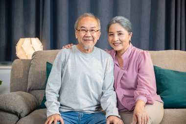 Mutlu Asyalı yaşlı çiftin portresi rahat bir koltukta otururken birini selamlıyor, yaşlı adam ve kadın evdeki bir kanepede yan yana oturuyor.