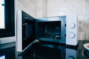 Zarif bir mutfak masasının üstünde modern teknoloji ve ev aletleri tasarımı sunan açık bir mikrodalga fırın kapısı. Modern mutfak ekipmanlarını sergilemek için mükemmel..