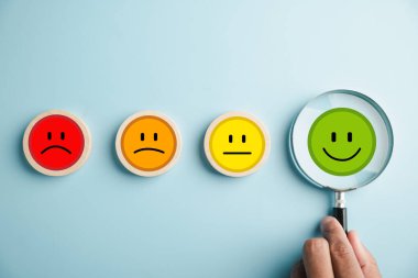 Eldeki büyüteç, duygular arasında seçilmiş mutlu gülen yüz simgesi. İyi geri bildirim notu, olumlu müşteri değerlendirmesi. Tecrübe, memnuniyet anketi, akıl sağlığı. Sertifika memnuniyeti gösterir.