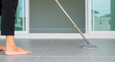 Kadın evin zeminini paspas ve bez parçasıyla temizliyor. Kendini temizliğe adamış bir ev hanımı. Profesyonel ev idaresi ve ev bakımı kavramlarını temsil eder