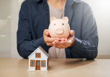 Bir domuz kumbarası ve ev modeli ile yakın çekim, tasarruf, finansal planlama ve emlak hedeflerini sembolize ediyor. Güvenli bir gelecek için ipotek seçenekleri ve ev sahipliği stratejilerini vurguluyor.