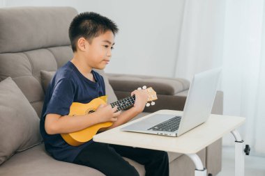 Çocuk kanepede ukulele öğrenirken dizüstü bilgisayarda özel ders izliyor. Konforlu oturma odası sahnesi, teknolojinin ve yaratıcı eğitimin modern ev ortamında entegrasyonunu vurguluyor..