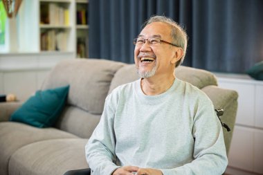 Asyalı yaşlı adamın yalnız portresi evdeki rahat oturma odasında rahat bir kanepede rahatlıyor, mutlu gülümseyen yaşlı bir erkek komik şakalara gülüyor ve iyi bir geleceği düşünüyor.