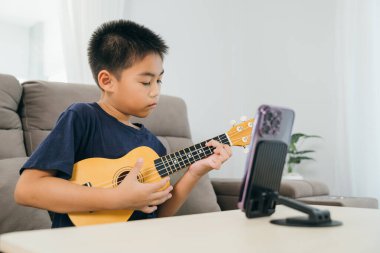 Akıllı telefon kullanarak müzik dersini canlı yayınlarken ukulele çalan Asyalı çocuk. Monte edilmiş telefon standı ve rahat oturma odası sahnesi modern öğrenme ve evde yaratıcı faaliyetler sergiliyor..