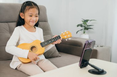 Genç bir kız ukulele çalan bir kanepede oturuyor, akıllı bir telefonla online müzik dersini yayınlıyor. Sahne, modern dijital araçlarla evde yaratıcı öğrenmenin altını çiziyor.