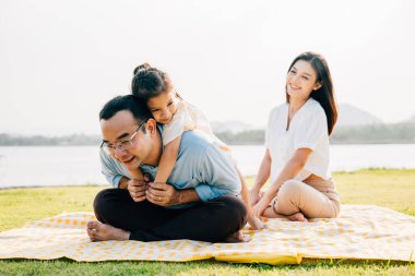 Piknik battaniyesinde oturan Asyalı aile parktaki çayır çimenlerinde oynuyorlar mutlu ebeveynler, çocuk babaya biniyor açık havada birlikte eğleniyorlar baba kızını taşıyor, aile ferahlığı.