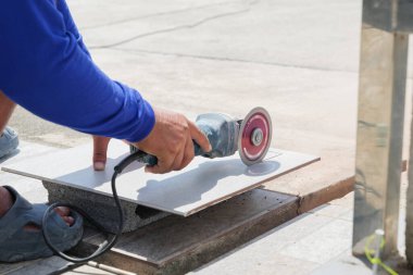 Açı öğütücü iş başında: mavi elbiseli bir işçi taştan bir yüzeydeki gri fayansı dikkatlice kesiyor, inşaat alanında uzman seramik kesme teknikleri sergiliyor.