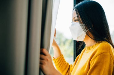 Tıbbi maskeli genç bir Asyalı kadın pencereden dışarı bakar COVID-19 yüzünden üzgün ve sıkıntılı hisseder. Korku izolasyonu ve salgınla ilgili kaygıları yansıtıyor..