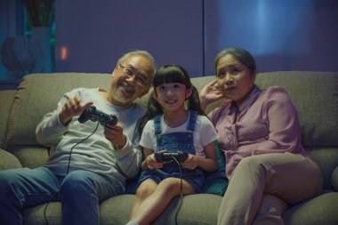 Konforlu bir kanepede video oyunu oynarken mutlu bir Asyalı aile bağı. Küçük bir kız, büyük anne ve büyükbabasıyla bilgisayar oyunu konsolu oynarken geceleri kanepede otururken kumandalı joysticklerle birlikte.