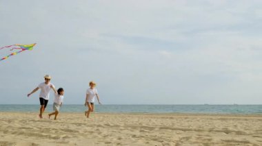 Asyalı bir aile sahilde koşarken gökyüzünde uçurtma uçuruyor. Mutlu ebeveynler, anne ve babalar ve çocukları uçurtmayla oynuyorlar, aileler birlikte vakit geçiriyorlar, yaz günü.