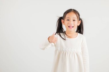 Beyaz arka planda izole edilmiş bir stüdyo portresindeki kendine güvenen Asyalı genç kız onay işareti yapıyor, anlaşma ve başarıyı simgeliyor. Anaokulu çocuk gülümsemesi pozitiflik ve memnuniyet yayar.
