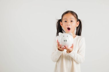 Asyalı küçük bir kız elinde domuz kumbarası, beyaz arka planda izole edilmiş stüdyoya bakıyor, mutlu bir anaokulu çocuk yaşam tarzı gülümsüyor, beyaz kumbara, Tayland kişisel para tasarrufu.