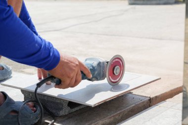 Mavi elbiseli bir işçinin gri bir fayans kesmek için değirmen kullandığı inşaat alanı sahnesi. Detaylı işçiliği ve fayans hazırlığını vurguluyor..