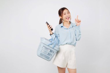 Heyecanlı Asyalı kadın, elinde alışveriş sepeti ve telefonuyla boş bir yeri işaret ediyor. Markette alışveriş keyfi yapıyor, seyyar market hayatını ve neşeli market tarzını gösteriyor. izole stüdyo beyaz arkaplan