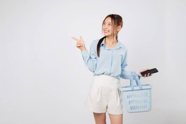 Gülümseyen Asyalı kadın elinde telefon ve alışveriş sepeti tutarken markette alışveriş yaşam tarzını mobil kolaylıkla gösteriyor. izole stüdyo beyaz arkaplan