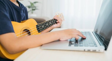 Kanepede otururken dizüstü bilgisayarı takip eden bir çocuğun ukulele üzerindeki parmaklarının yakın çekimi. Bu ayrıntı onun çevrimiçi eğitim yoluyla bir evde müzik öğrenmeye odaklanmasını sağlıyor..