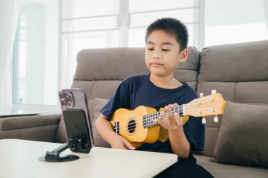 Genç bir çocuk kanepede ukulele çalıyor ve önünde müzik dersleri için bir akıllı telefon var. Evde öğrenim kavramı, yaratıcı eğitimde modern öğretim araçlarını vurguluyor.